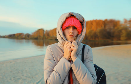 Woman is cold, freezing trying to warm up outdoors on the beach in autumn day, standing on sea coast, wearing warm jacket with hood, hatの写真素材