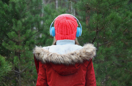 Back view modern woman enjoying listening to music with headphones in hat, jacket in winter forest against Christmas tree background with snowの写真素材