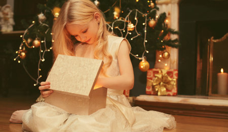 Happy little girl child opens festive gift box sitting near Christmas tree and fireplace with holiday lights at homeの写真素材