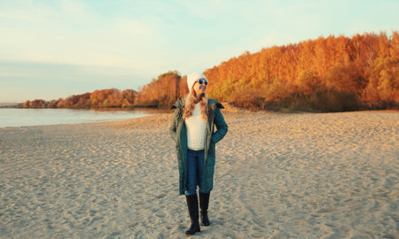 Traveler stylish woman full length standing on sunny beach looking away, enjoying amazing landscape, warm weather, wear hat, jacket, sea coast, nature background, trees with yellow leavesの写真素材