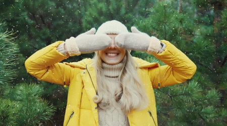 Happy smiling young woman having fun enjoys snow, warm weather in winter park against Christmas tree, joyful girl in hat with mittens standing in snowy forest with snowflakes, festive backgroundの写真素材