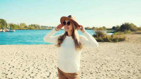 Stylish happy woman standing on sunny beach, warm weather, smiles, looking away, wear hat, glasses, white sweater, on sea coast backgroundの写真素材