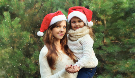 Family, celebration, holiday. Happy mother hugging her little girl child in santa hat together near Christmas tree in forest with snowの写真素材