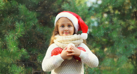 Christmas portrait of happy smiling little girl child in santa red hat with ball toy decoration outdoors on green tree background with snowの写真素材