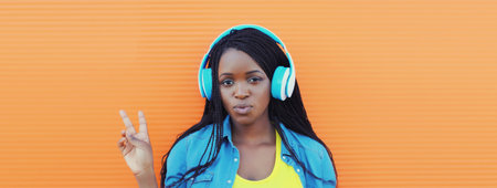 Portrait of happy African young woman listening to music with headphones, cool modern black teenage girl smiles on colorful bright orange background, summer dayの写真素材
