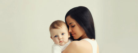 Happy young mother holding her cute baby sitting together in white room at homeの写真素材