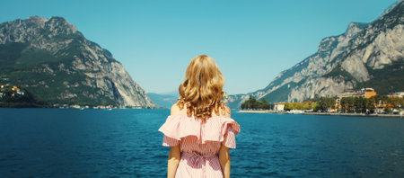 Summer vacation, beautiful young woman tourist standing on lake in Italy enjoying amazing sea landscape, mountains backgroundの写真素材