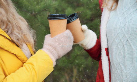 Close up two women friends, female hands holding cups of hot coffee drink against Christmas trees in...