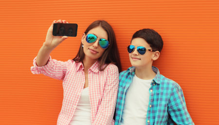 Happy mother with son teenager taking selfie with smartphone wearing summer glasses in the city on orange wall backgroundの写真素材