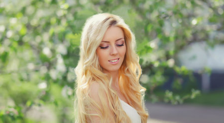 Beautiful young woman with blonde long hair in spring blooming garden on a sunny day against flowers trees backgroundの写真素材
