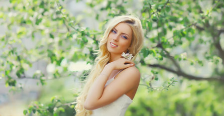 Beautiful young woman with blonde long hair in spring blooming garden on a sunny day against flowers trees backgroundの写真素材
