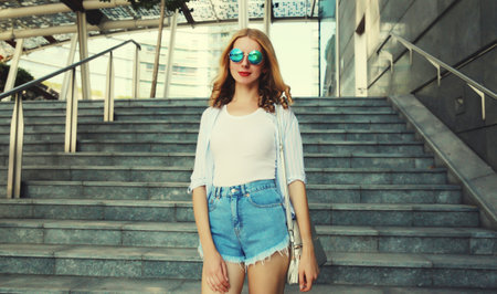 Beautiful modern young woman looking at camera standing on city street in summer wearing shorts, shirt, glassesの写真素材