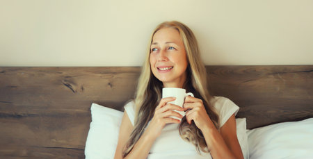 Happy smiling mature woman enjoying with cup of hot coffee lying on bed at home in the morningの写真素材