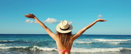 Summer vacation, rear view of beautiful happy slim woman in bikini swimsuit and tourist straw hat raising her hands up on the beach on sunny day on sea coast with wavesの写真素材