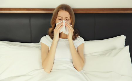 Sick exhausted young woman sneezing blow nose using tissue, unhealthy suffering girl feels unwell lying on bed at homeの写真素材