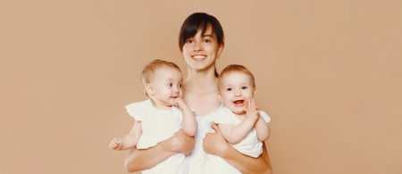Portrait of happy smiling young mother holding twin babies on brown studio backgroundの写真素材