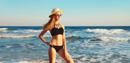 Summer vacation, beautiful happy slim woman in black bikini swimsuit, tourist hat looking away standing on the beach on sunny sea coast background with waves and blue skyの写真素材