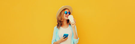 Summer stylish happy young woman drinking coffee with phone, cool girl using smartphone holding cup of coffee dressed tourist straw hat looking away standing on yellow backgroundの写真素材