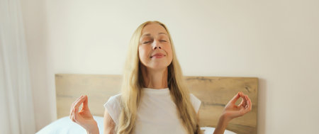 Calm relaxed healthy young woman meditates, girl practicing doing yoga exercises sitting in lotus pose on white bed at home, stress reliefの写真素材