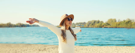 Happy smiling woman traveler raising her hands up on beach, enjoying warm weather, wear hat, white sweater, standing on sea coast, blue sky, nature backgroundの写真素材