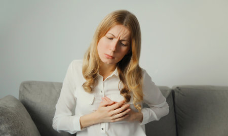 Unhealthy young woman touching hands her chest suffering from heart pain, feeling bad, heart attack, having health problems, sitting on the couch at homeの写真素材