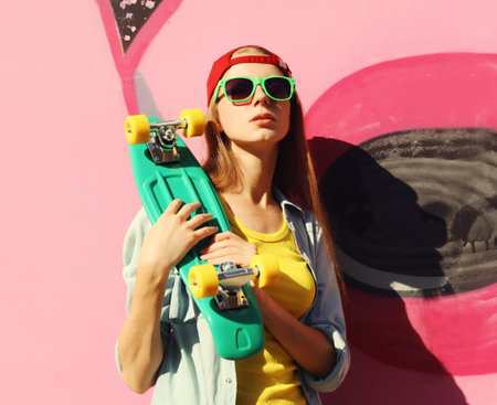 Portrait of stylish young girl with skateboard in glasses, modern teenager girl posing against the background of colorful pink graffiti wallの写真素材