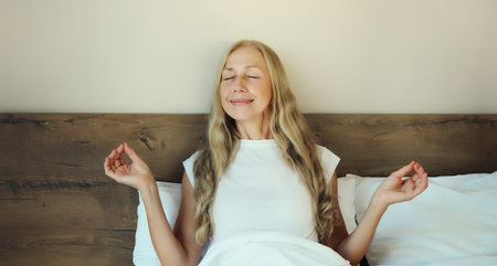 Calm relaxed healthy young woman meditates, girl practicing doing yoga exercises sitting in lotus pose on white bed at home in the morning, stress reliefの写真素材
