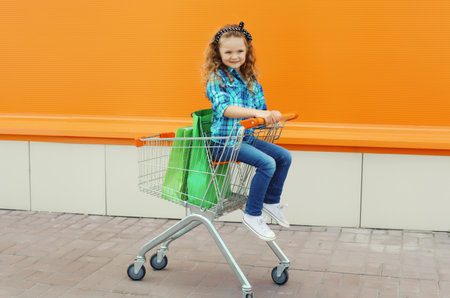Happy little girl child sitting in trolley cart and colorful shopping bags, kid on city streetの写真素材