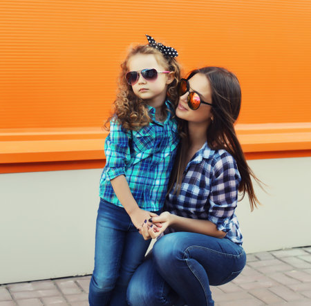 Beautiful stylish mother and child daughter together, happy mom with kid girl in glasses, casual clothing in the city on an orange backgroundの写真素材