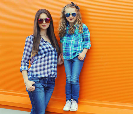 Beautiful stylish mother and child daughter together, happy mom with kid girl in glasses, casual clothing in the city on an orange backgroundの写真素材
