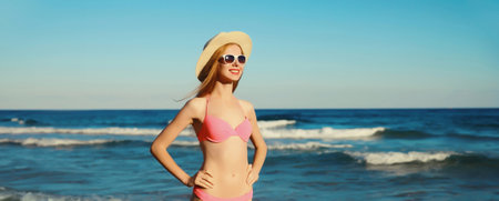 Summer vacation, beautiful happy slim woman in bikini swimsuit, tourist hat looking away standing on the beach on sunny sea coast background with wavesの写真素材