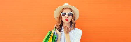 Stylish beautiful young woman model with shopping bags blowing a kiss in summer straw hat on orange studio backgroundの写真素材
