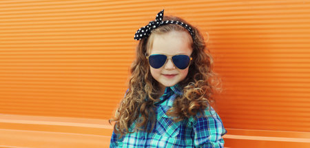 Happy kid girl with curly hair, stylish little child posing on city street on orange wall backgroundの写真素材
