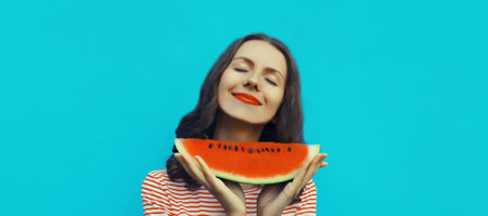 Summer portrait of happy young woman enjoying eating fresh slice of watermelon fruit on blue studio backgroundの写真素材
