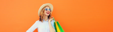 Fashion, purchase. Portrait of stylish beautiful happy young woman posing with colorful shopping bags, girl in summer hat standing on bright orange backgroundの写真素材