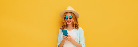 Summer portrait of happy traveler young woman with mobile phone looking at smartphone wearing a straw tourist hat, casual clothing on yellow studio backgroundの写真素材