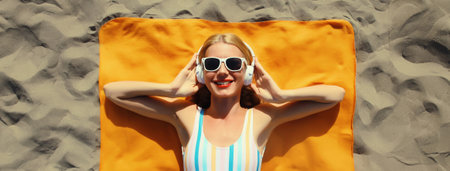 Summer vacation, tourism, happy cheerful young woman listening to music lying on sand on the beach, relaxed tourist girl in headphones on towel in swimsuit, glasses, sunny day, top viewの写真素材