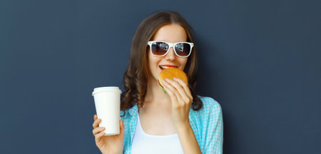 Portrait of happy cheerful young woman eating burger fast food holding cup of coffee, teenage girl having fun isolated on black wall studio backgroundの写真素材