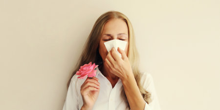 Sick exhausted woman suffering from pollen holds flower sneezing blow nose using tissue on white studio backgroundの写真素材