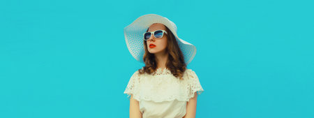 Beautiful stylish young woman, fashionable female model in summer white straw hat posing on blue wall background, looking awayの写真素材