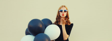 Portrait of beautiful happy young woman celebrating with bunch of black and white balloons in summer dress and blowing her lips kiss on white backgroundの写真素材