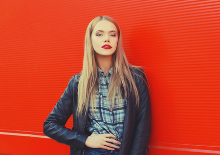 Fashionable portrait of beautiful blonde young woman posing in black leather jacket on red backgroundの写真素材
