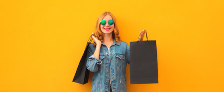 Shopping day, stylish beautiful happy smiling young woman posing with black shopping bags wearing denim clothing on yellow studio backgroundの写真素材