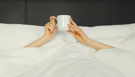 Cheerful lazy woman waking up after sleeping in the morning while lying in white soft comfortable bed showing empty cup coffee stretching her hands up from under the blanket in bedroom at homeの写真素材