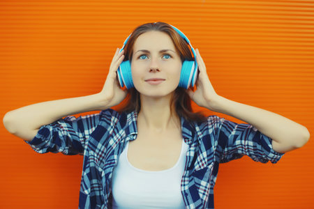 Summer portrait of happy modern young woman listening to music with headphones on orange wall...