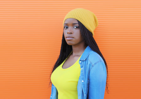 Fashionable portrait of young African woman in casual clothing, stylish modern black girl in yellow hat with dreadlocks posing in the city on bright orange wall backgroundの写真素材