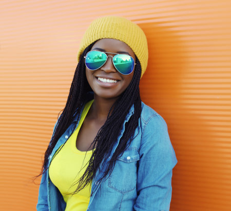 Portrait of happy stylish young African woman in casual clothing, glasses with dreadlocks, black girl smiling wear yellow hat posing in the city against a vivid orange wall backgroundの写真素材