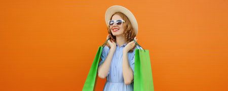 Fashion, purchase. Portrait of stylish beautiful happy young woman posing with colorful green shopping bags, girl smiles wear summer dress standing on studio orange wall backgroundの写真素材