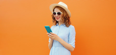 Summer portrait of happy traveler young woman with mobile phone, girl looking at smartphone wearing a straw tourist hat, casual clothing on orange studio backgroundの写真素材