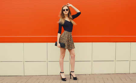 Beautiful stylish blonde young woman full length in leopard print skirt in heels with black handbag clutch standing on red wall, city streetの写真素材